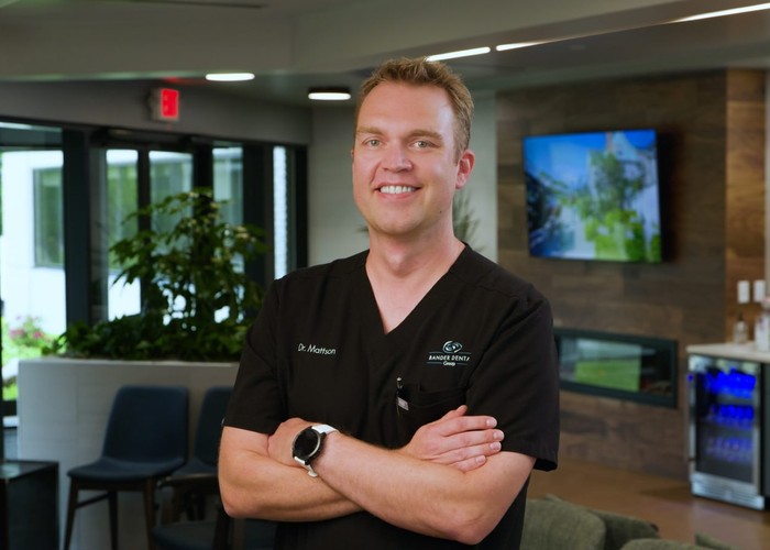 A man in a blue plaid shirt smiles warmly at the camera, with a blurred modern office interior in the background.